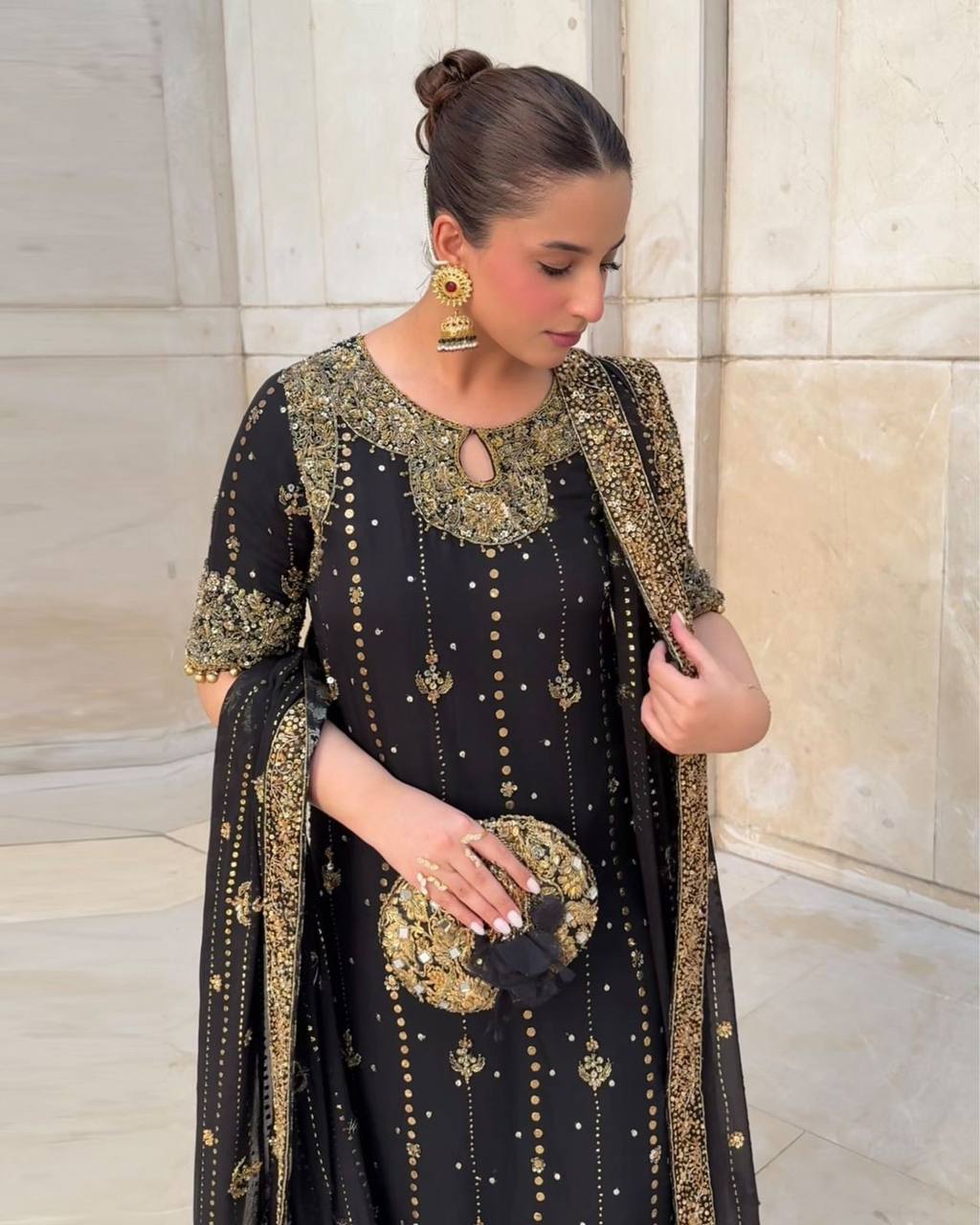 Woman wearing a black and gold traditional outfit with intricate designs, standing against a marble wall.