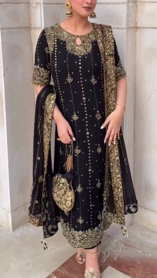 Woman wearing a black and gold traditional outfit with embroidery, standing against a light-colored wall.