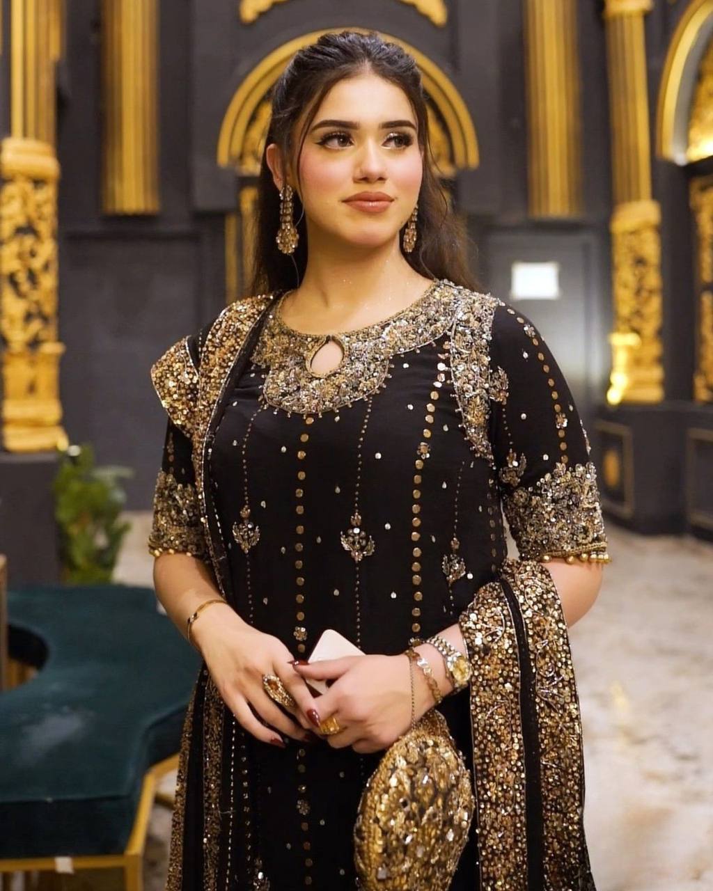 Woman in a black and gold traditional outfit with intricate designs in an ornate indoor setting.