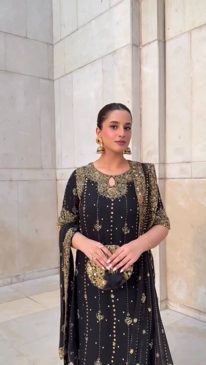 Woman in a black and gold traditional outfit standing against a light stone wall.