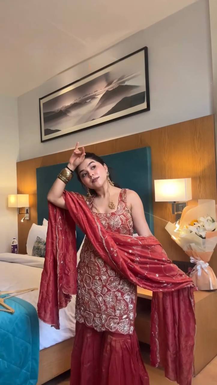 Woman in a red traditional outfit posing in a room with a bed and lamp.